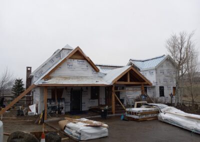 A work in progress: residential construction site with a partially completed house featuring exposed tyvek house wrap and roofing underlayment, surrounded by building materials and scaffolding on an overcast day.