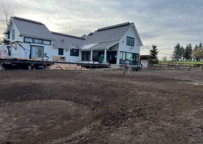 A large modern farmhouse with white siding and metal roofs undergoing landscaping work with freshly tilled soil in the foreground.