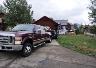 A maroon pickup truck hauling a flatbed trailer loaded with construction supplies parked in front of a wooden house with overcast skies above.