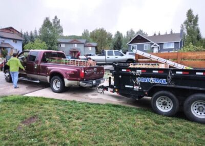 A maroon pickup truck towing a flatbed utility trailer parked on a residential street with a person walking towards the house and another vehicle parked in the background.